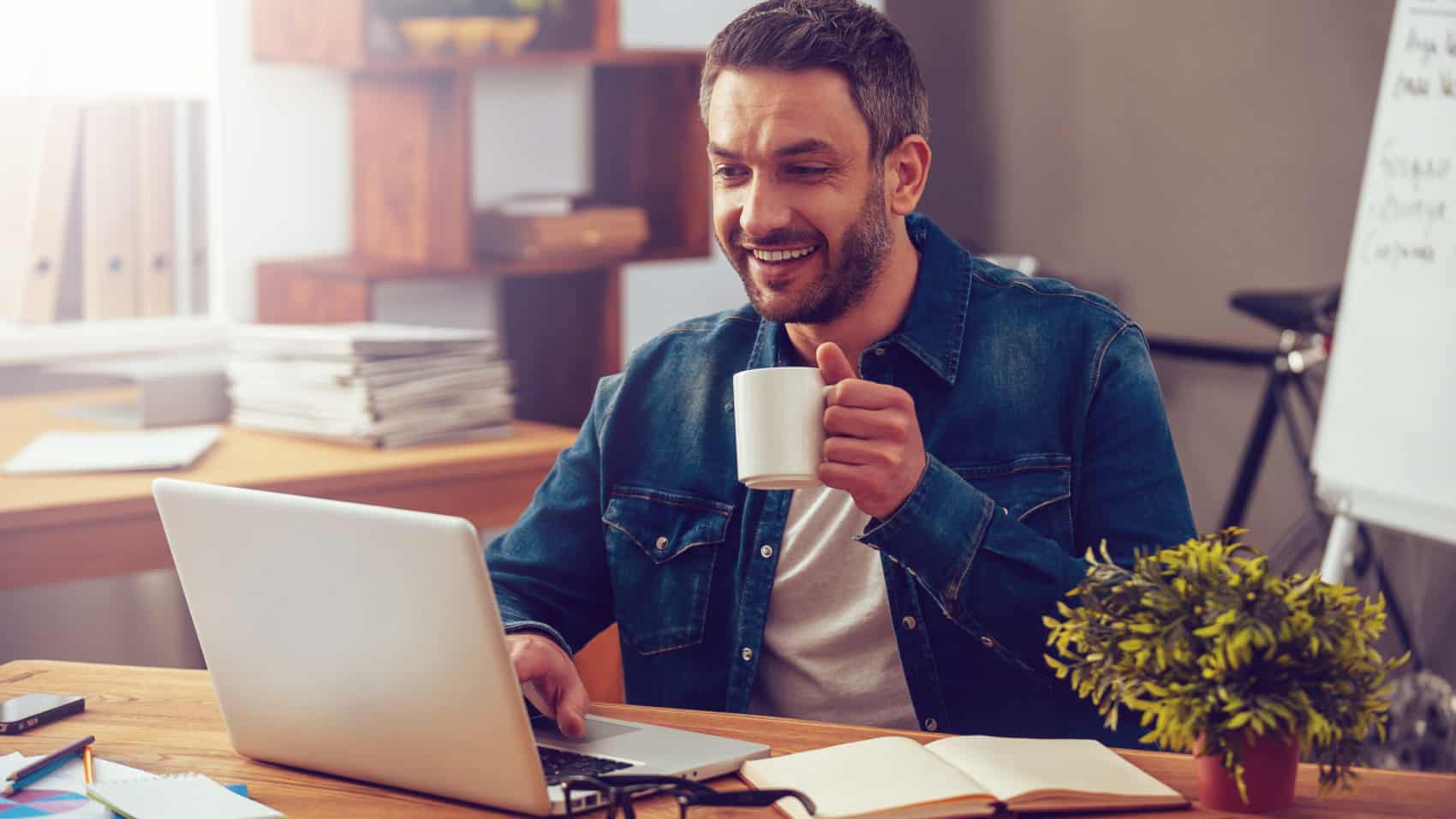 Confident young man working on laptop and holding coffee cup while sitting at his working place in office